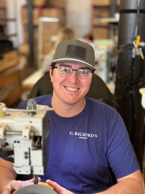 Tim, stitcher at C. Richard's, wearing a branded leather patch cap and smiling at his sewing machine